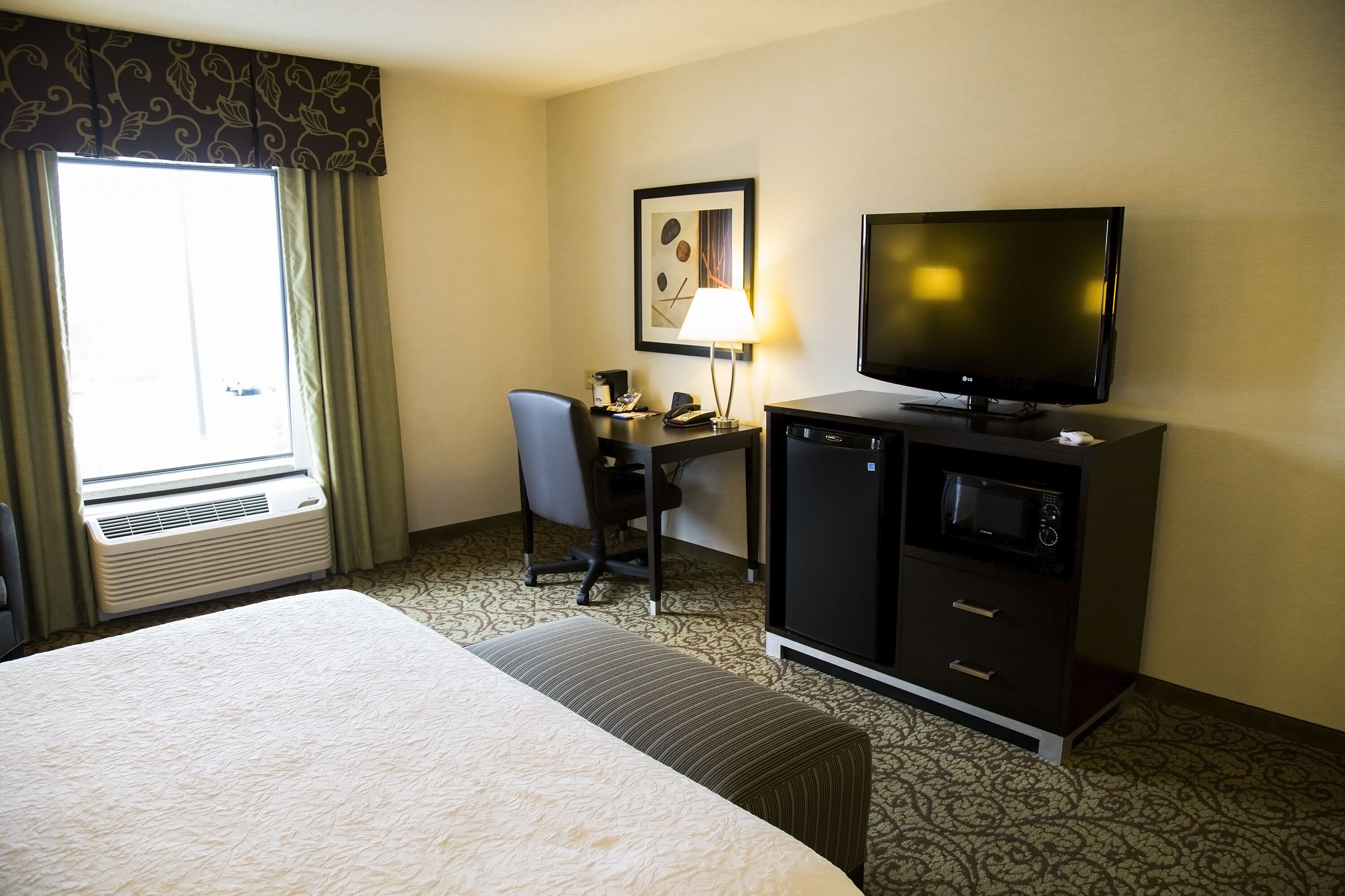 A hotel room with a bed, a desk with a chair, a television, a microwave, and a window with curtains.