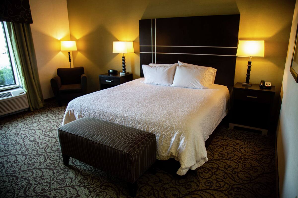 Quarto de hotel com uma cama grande, duas mesas de cabeceira com luminárias, uma cadeira e um tapete estampado.
