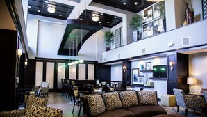Eine geräumige Hotelhalle mit hoher Decke, eine Sitzecke mit gemusterten Sofas, ein Essbereich mit Tischen und Stühlen sowie ein an der Wand befestigter Fernseher.