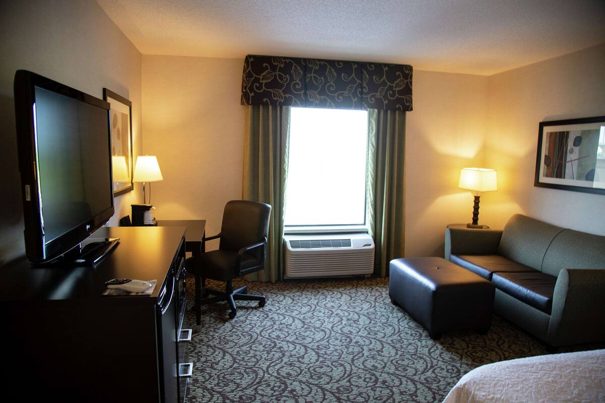 Quarto de hotel com televisão de tela plana, escrivaninha com cadeira, sofá, banquinho e uma grande janela com cortinas.