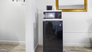 Un refrigerador compacto con un microondas encima, ubicado en una esquina de una habitación con paredes blancas y piso de madera.