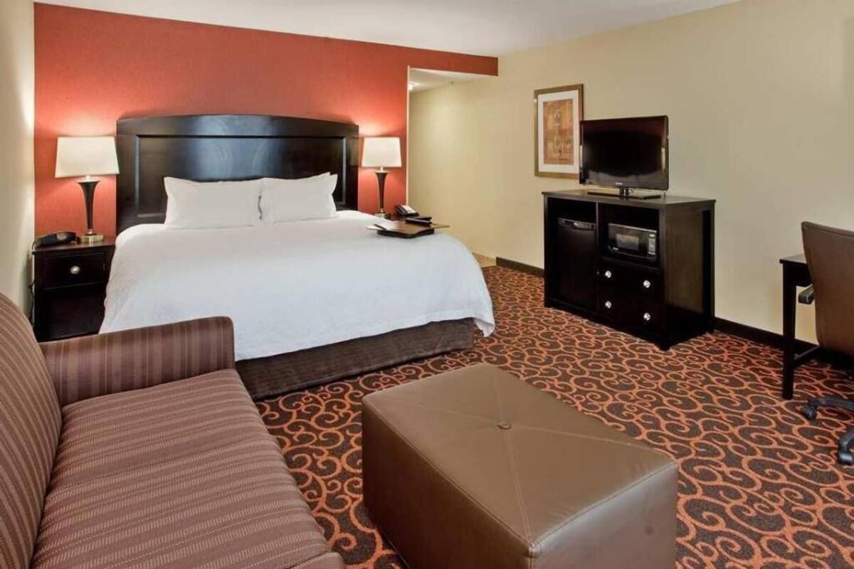 Una habitación de hotel con cama, sofá, otoman, televisor y escritorio.