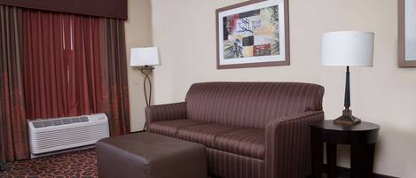 A hotel room with a striped sofa, an ottoman, a standing lamp, a framed abstract painting, and a wall-mounted air conditioner.