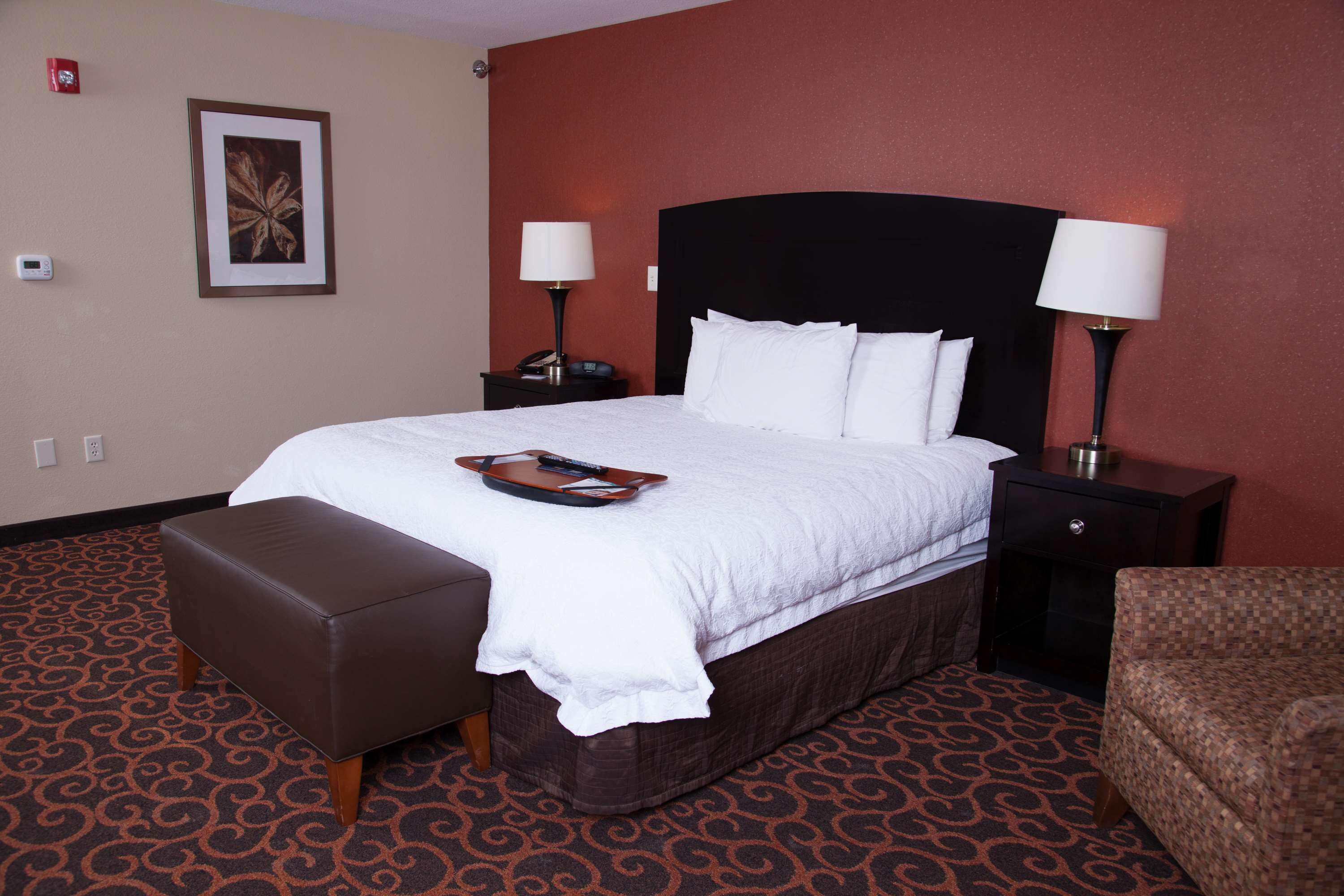 Una habitación de hotel con una cama grande, dos mesitas de noche con lámparas, un reposapiés marrón y una alfombra estampada.