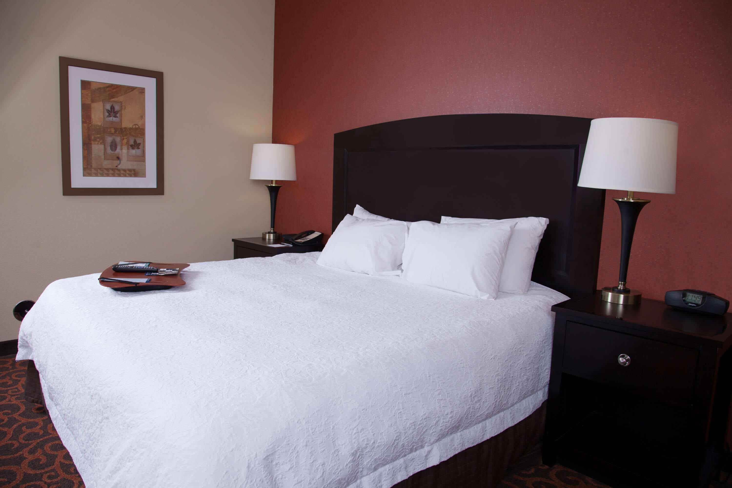 A neatly made bed with white linens, a remote control, and a smartphone on the side table. There are two bedside lamps and a framed picture on the wall.