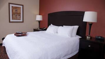 A neatly made bed with white linens, a remote control, and a smartphone on the side table. There are two bedside lamps and a framed picture on the wall.