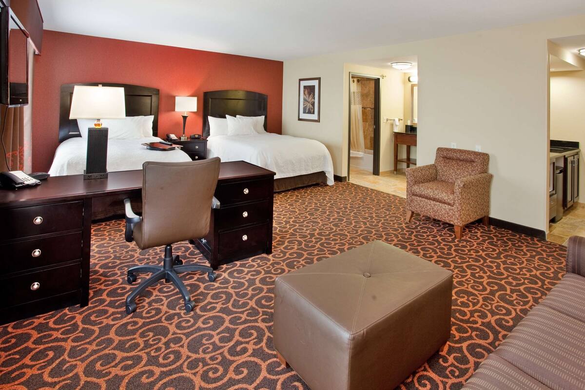 Habitación de hotel con cama, escritorio, silla, reposapiés y baño visible a través de una puerta abierta.