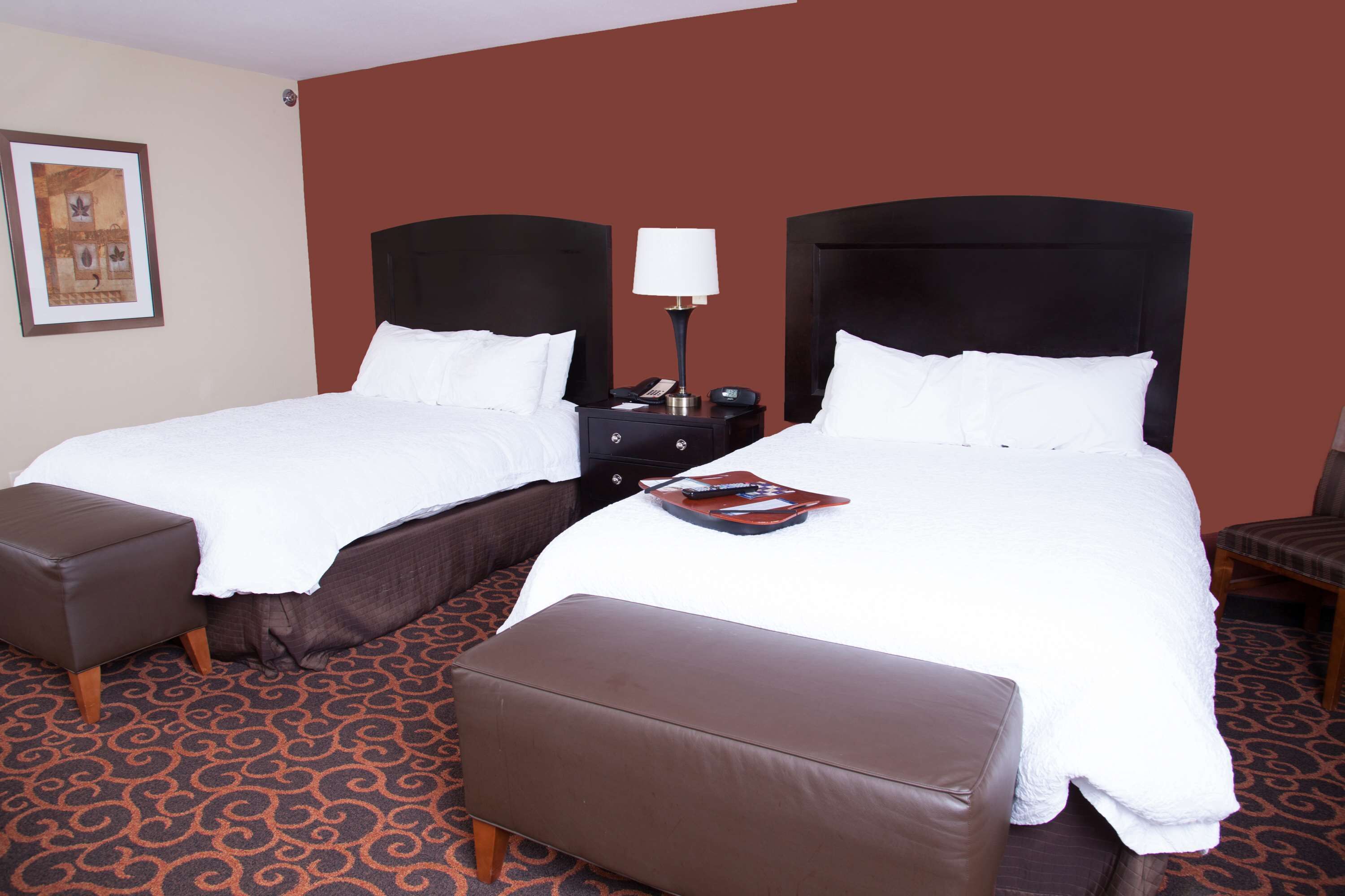 A hotel room with two beds, a nightstand with a lamp, and a brown ottoman.