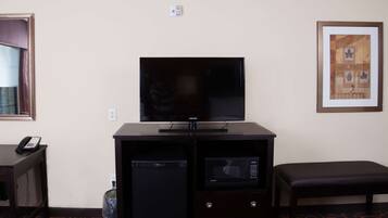A hotel room with a flat-screen TV on a wooden cabinet, a small refrigerator, a microwave, a telephone, and a framed picture on the wall.