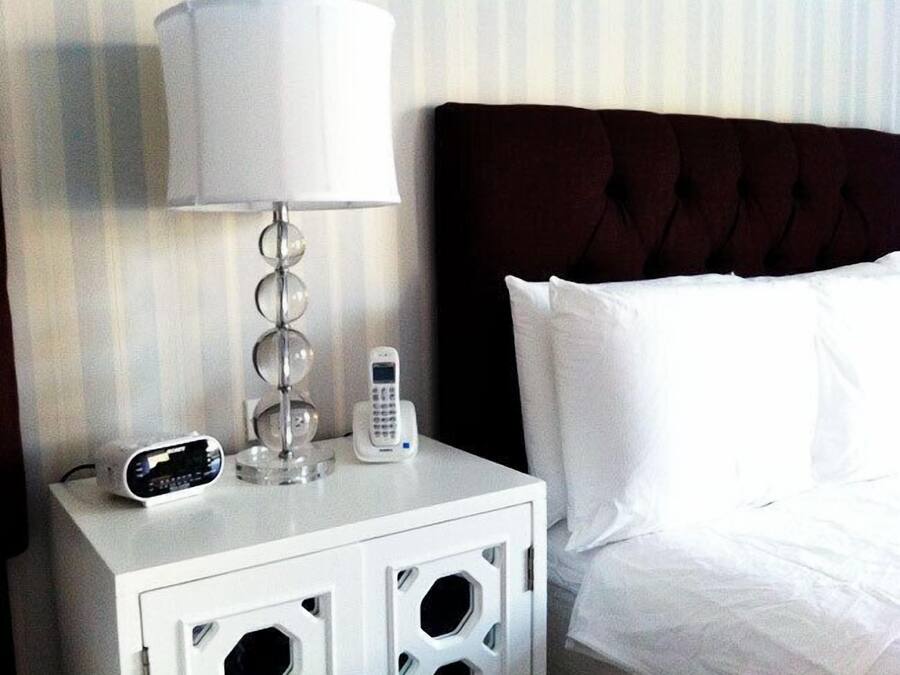 Ein ordentlich bezogenes Bett mit weißen Leinen, ein weißer Nachttisch mit Lampe und Telefon.