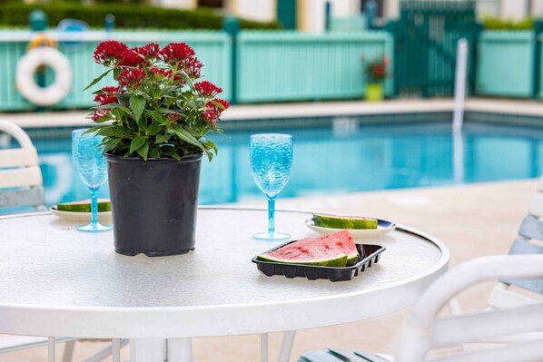 Une table placée au bord de la piscine, avec un pot de fleurs, deux verres, une tranche de pastèque et un plateau.