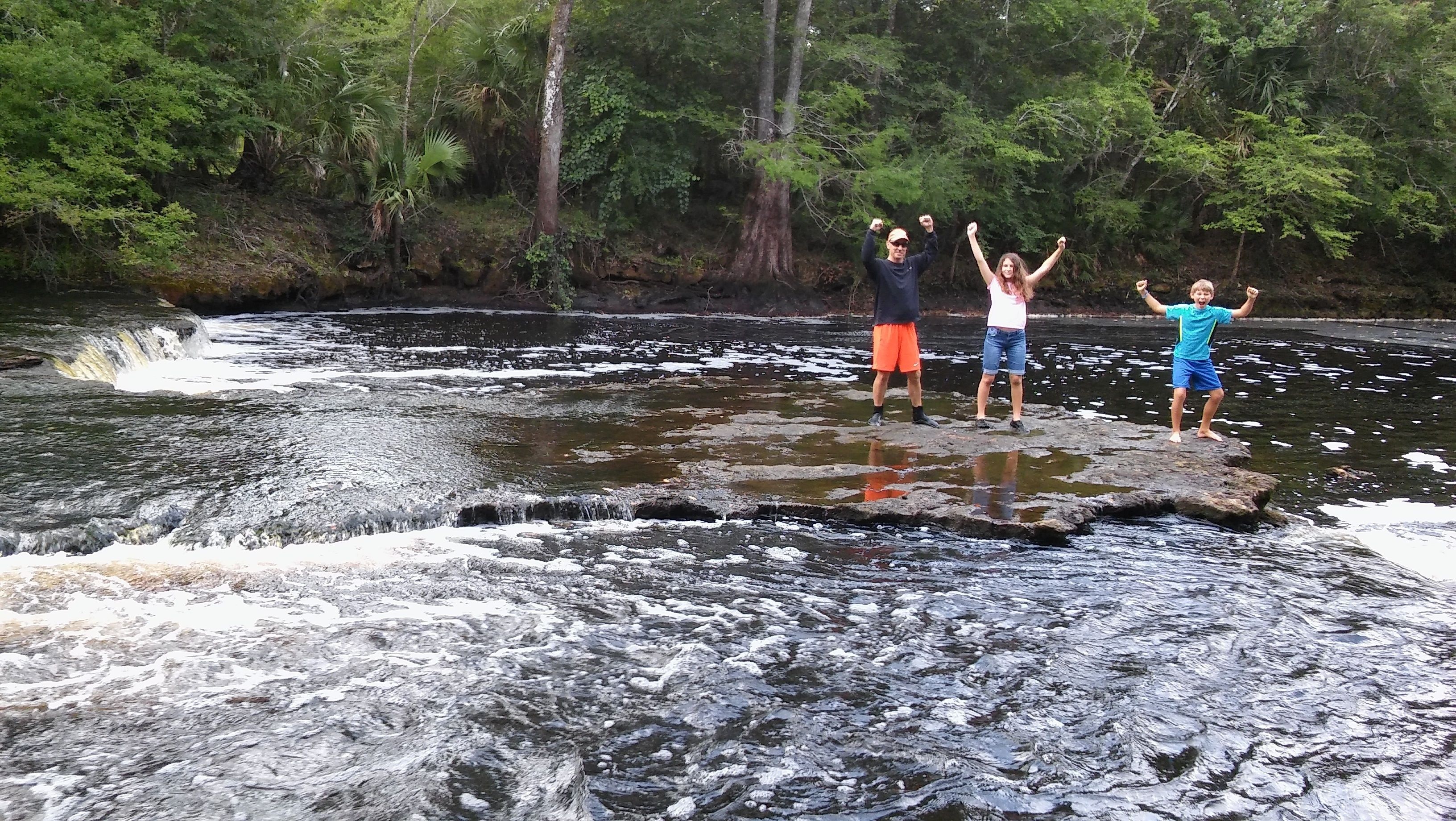 A group of people standing on rocks by a flowing river surrounded by trees.