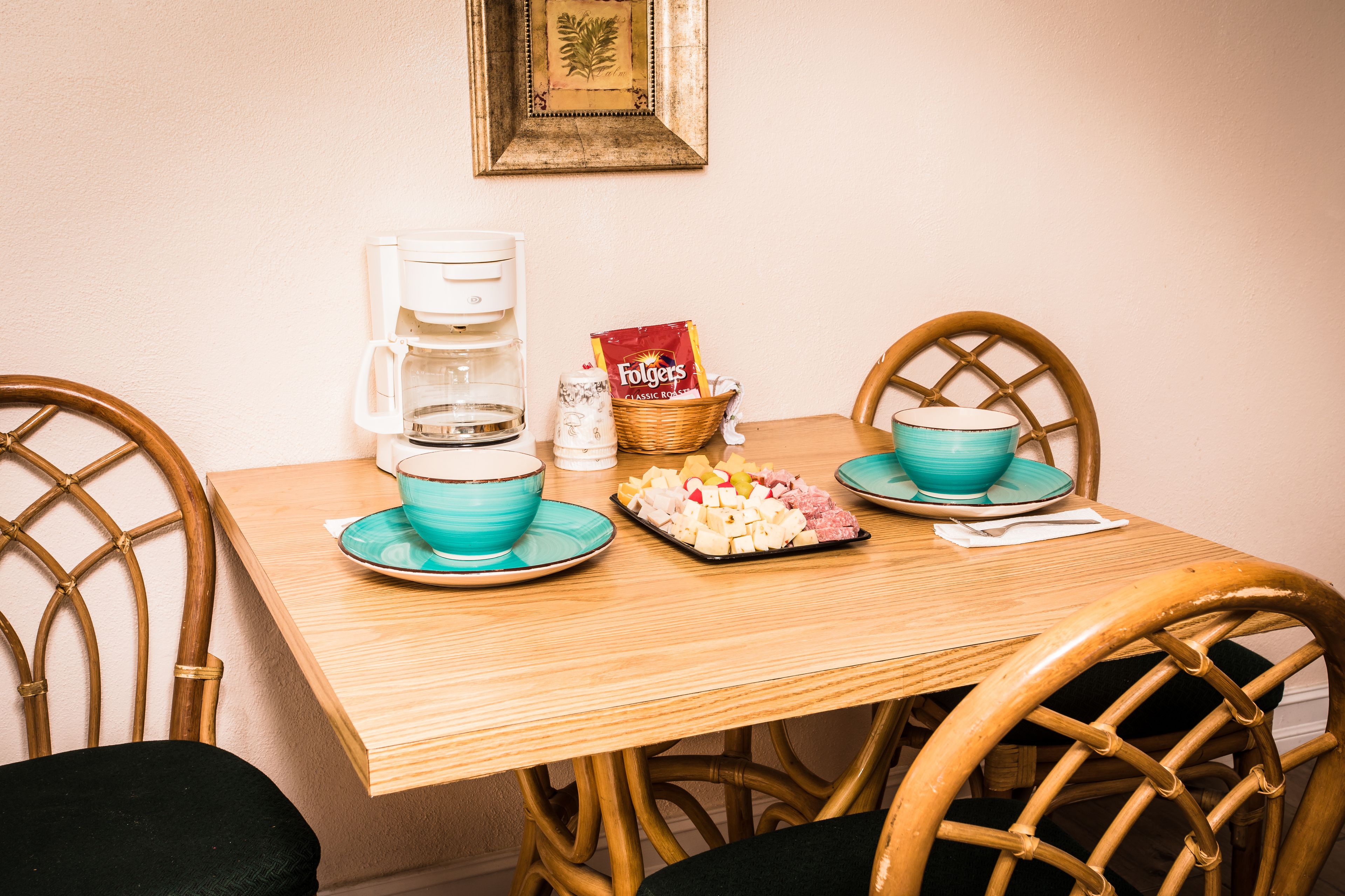 Une table en bois avec une machine à café, deux bols turquoise, une assiette de fromage et un panier de collations.