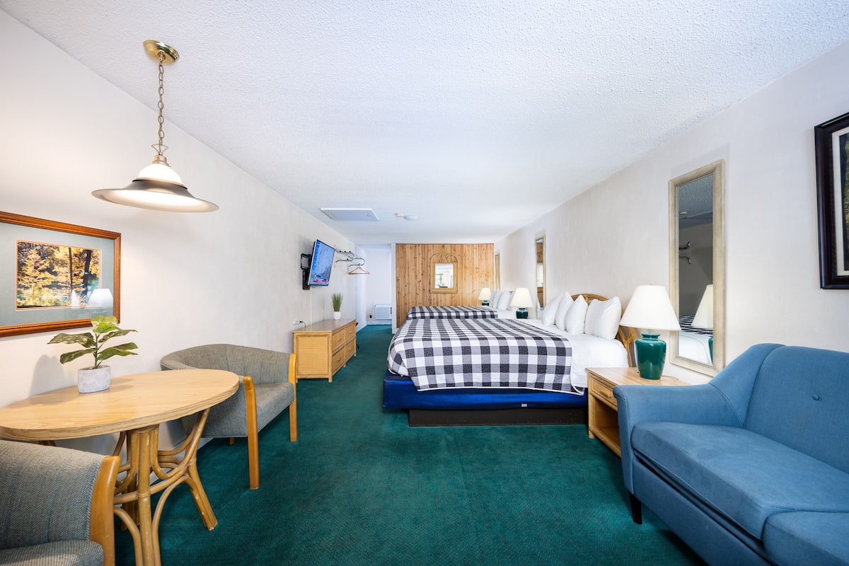 Habitación de hotel con cama, sofá azul, mesa de madera, una silla, televisor y un cuadro en la pared.