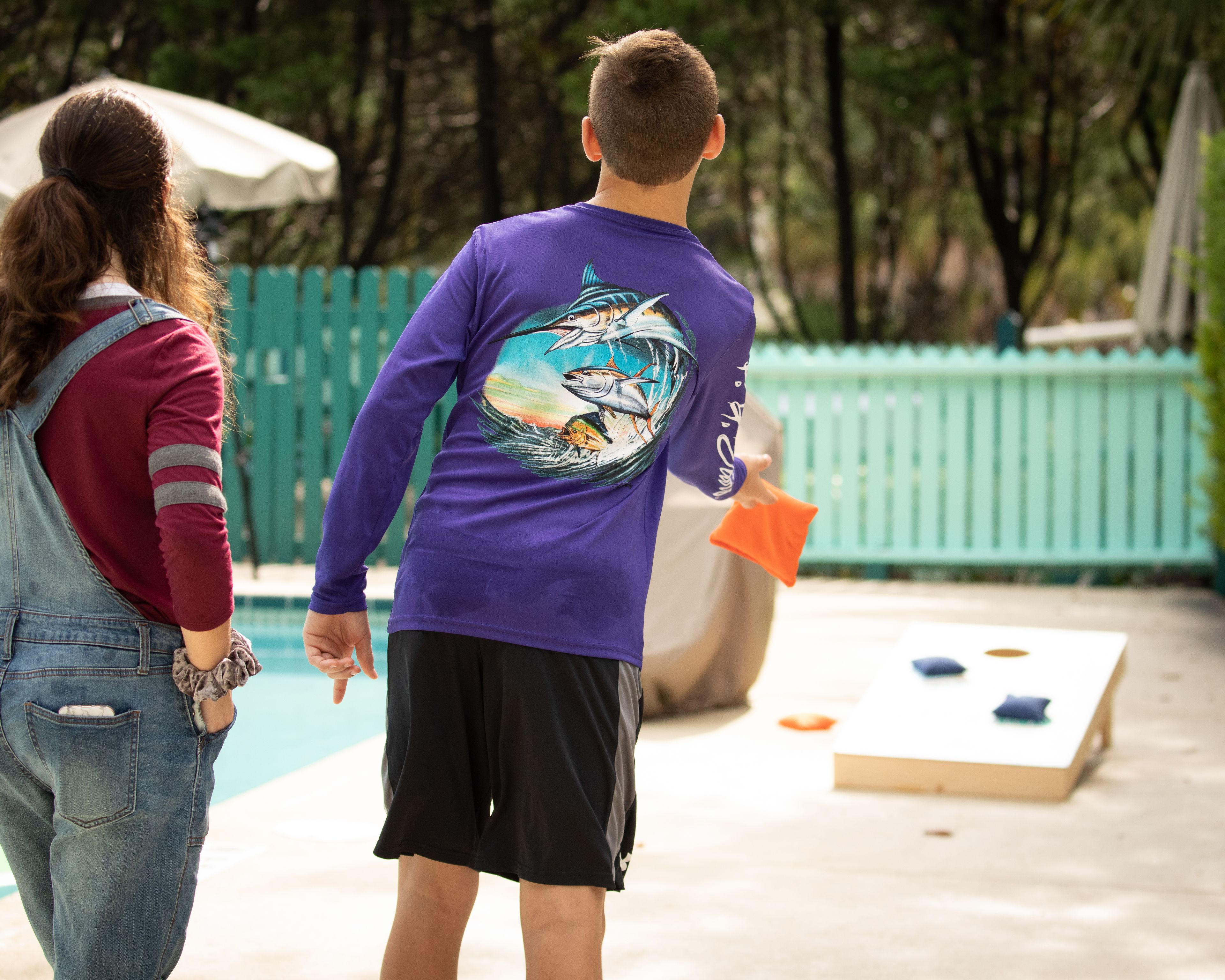 A person in a purple shirt with a graphic design, holding an orange object, standing near a pool area with a green fence and a white umbrella in the background.