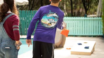 A person in a purple shirt with a graphic design, holding an orange object, standing near a pool area with a green fence and a white umbrella in the background.