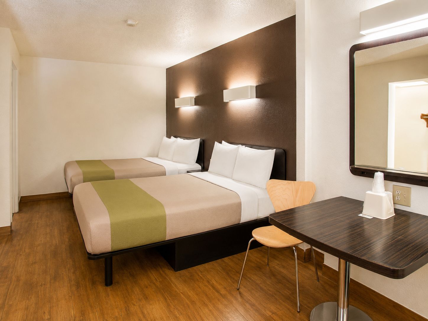 A hotel room with two beds, a wooden floor, a desk with a chair, and a large mirror.