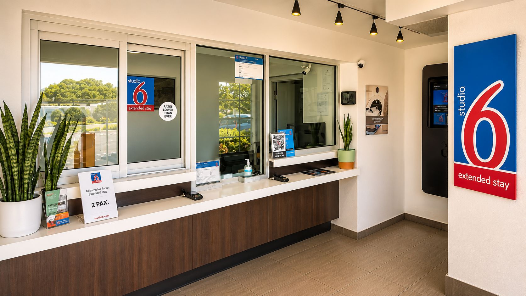 A hotel reception desk with a digital screen, a plant, and a sign indicating extended stay rates.