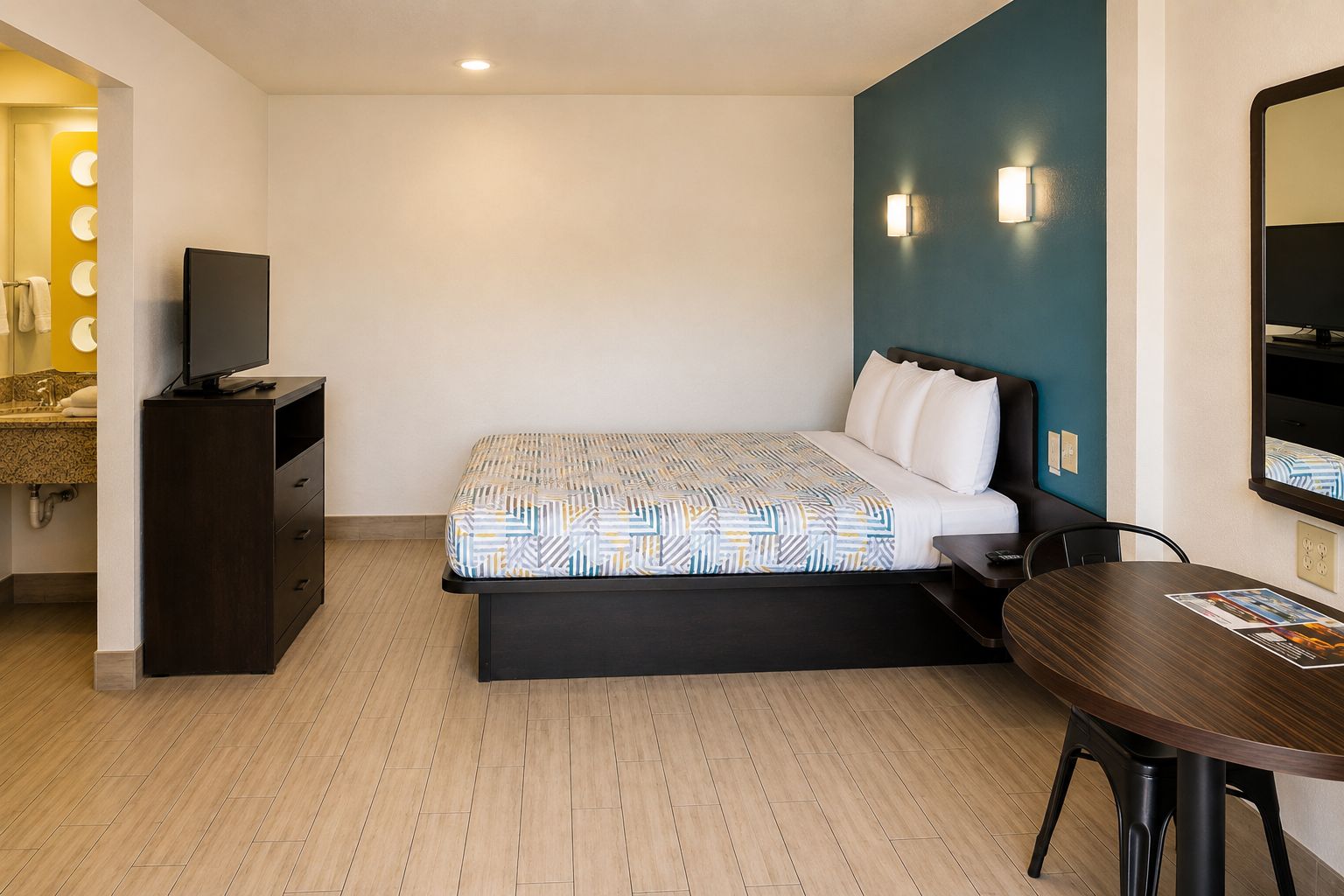 A hotel room with a bed, a desk, a chair, a TV, and a bathroom with a mirror and lights.
