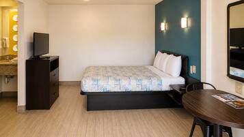 A hotel room with a bed, a desk, a chair, a TV, and a bathroom with a mirror and lights.