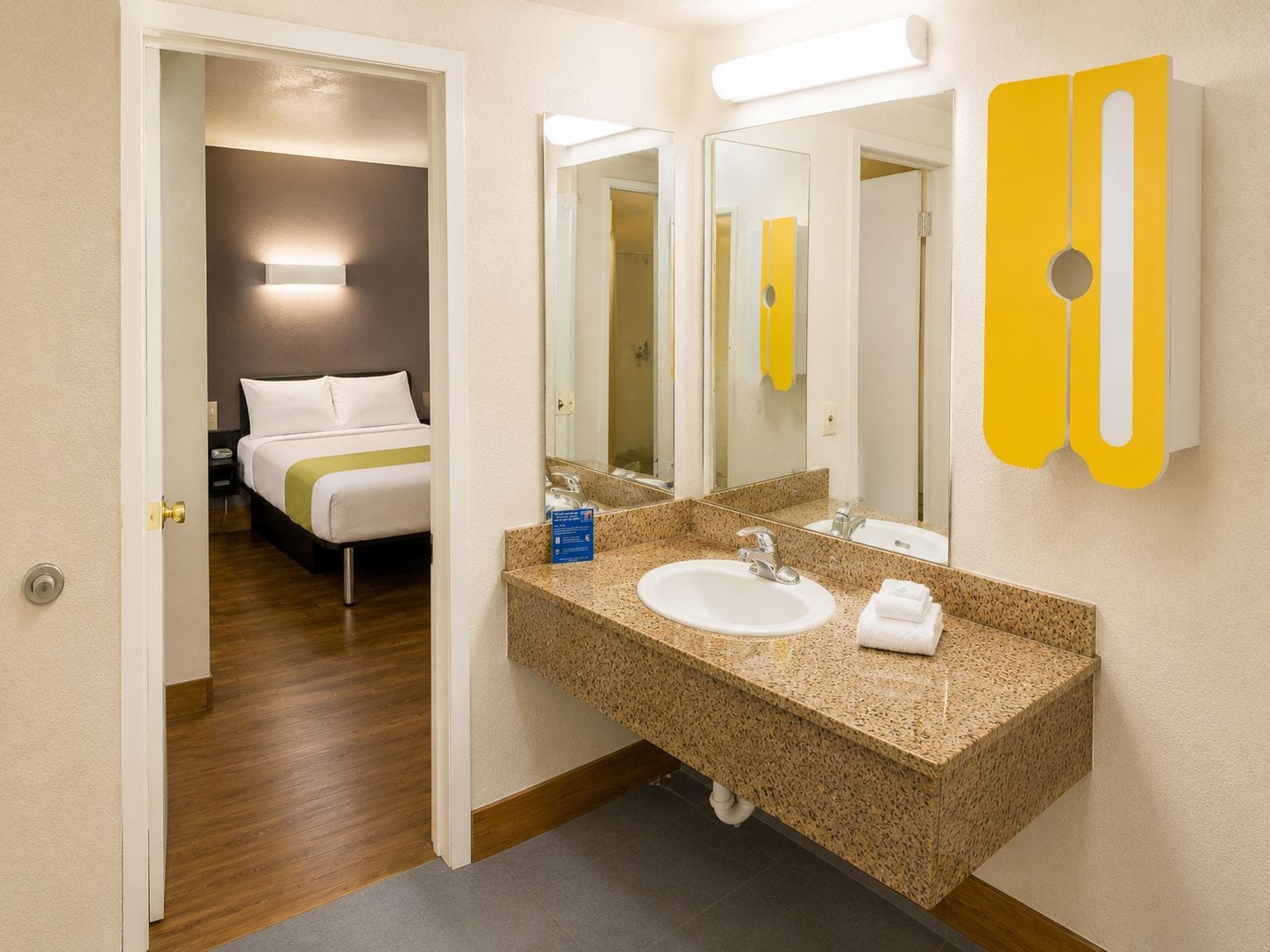 Baño de hotel con lavamanos de encimera de granito, un espejo y una cama visible a través de una puerta abierta.