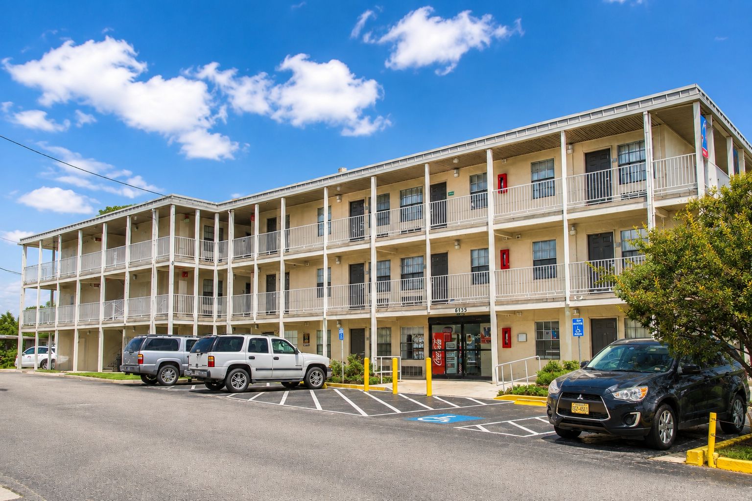 A multi-story motel with a parking lot, several vehicles parked, and a Coca-Cola sign.