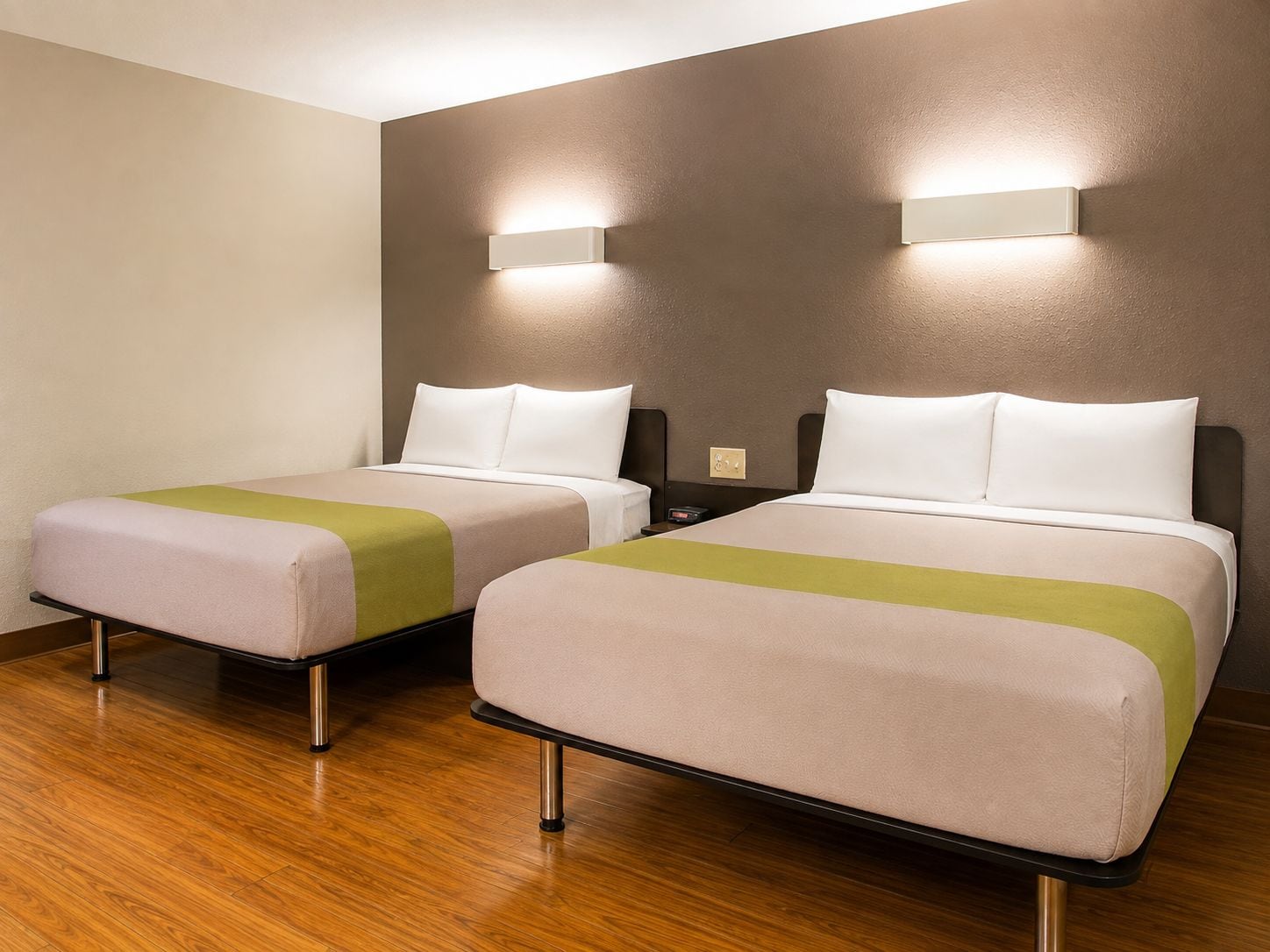 A hotel room with two beds, each with a pillow and a blanket. There is a wall-mounted light fixture above each bed.