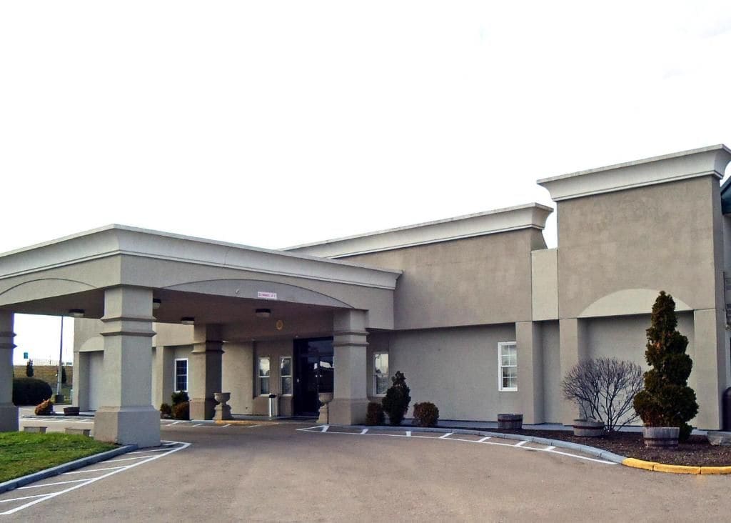 A hotel entrance with a covered driveway, a parking lot, and a landscaped area with shrubs and a tree.