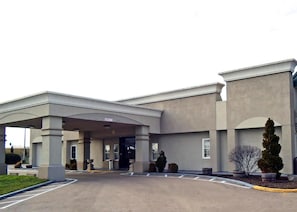A hotel entrance with a covered driveway, a parking lot, and a landscaped area with shrubs and a tree. - Motel 6 Troy, OH (Troy)