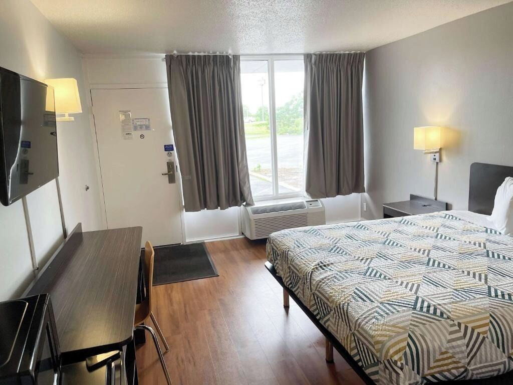 Una habitación de hotel con cama, escritorio, silla, lámpara, ventana con cortinas y unidad de aire acondicionado.