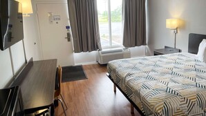 A hotel room with a bed, a desk, a chair, a TV, a window with curtains, and a wall-mounted lamp. - Motel 6 Troy, OH (Troy)