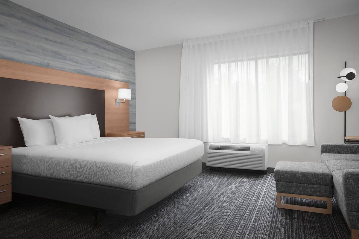 Une chambre d’hôtel moderne dotée d’un grand lit, d’une table de chevet, d’une chaise, d’une fenêtre avec des rideaux et d’un climatiseur fixé au mur.