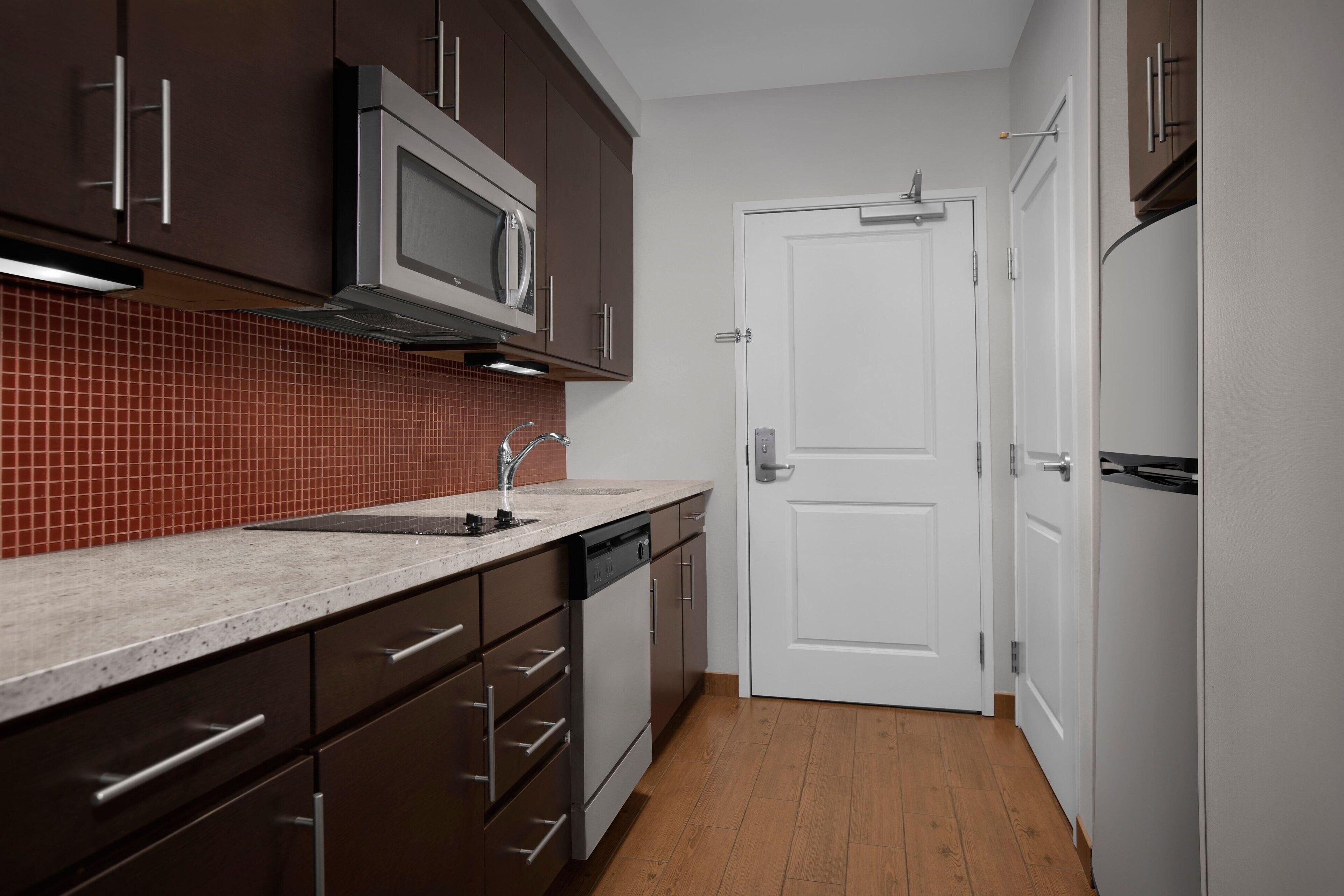 A modern kitchen with dark cabinets, a stainless steel microwave, a dishwasher, and a white door.