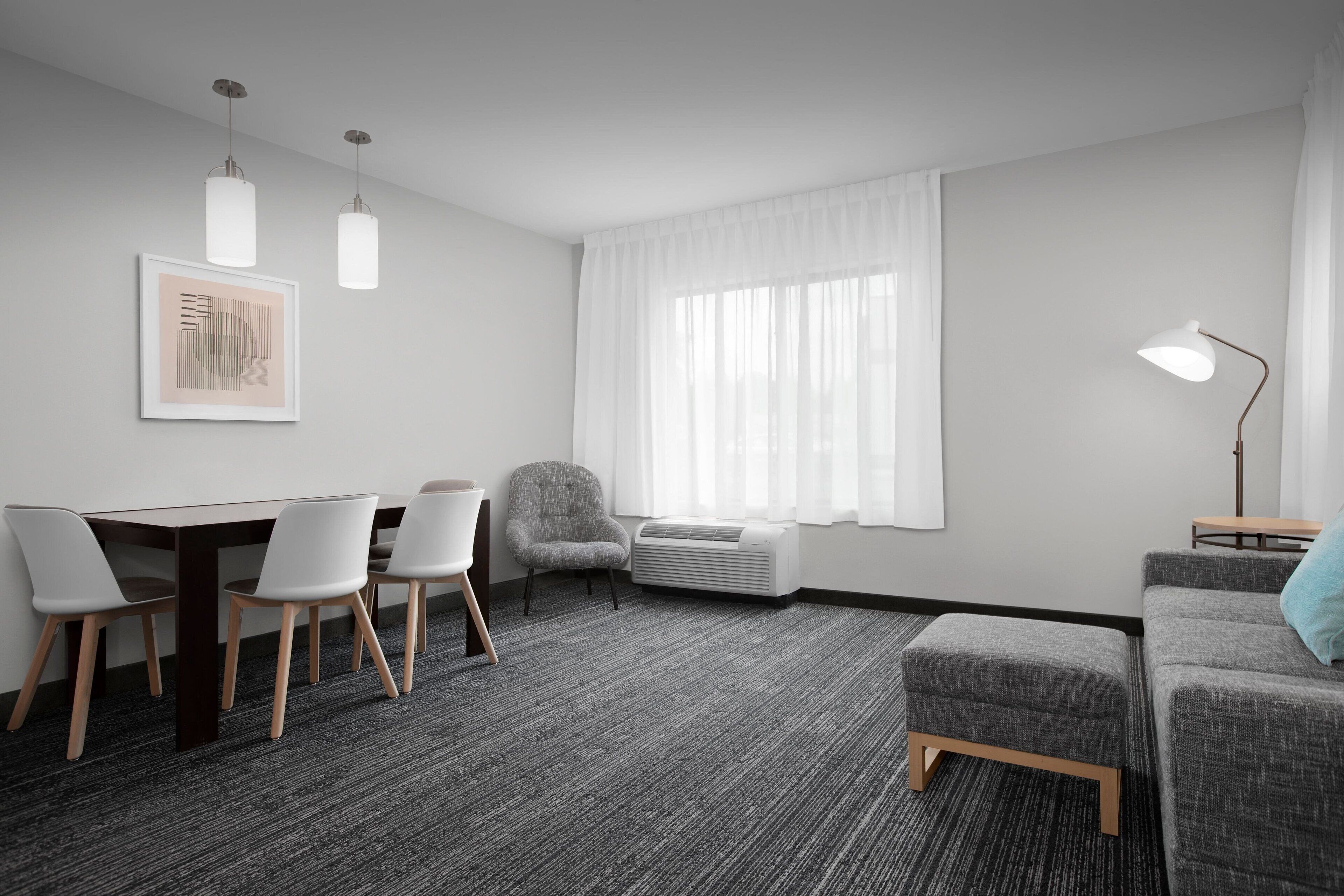 A modern hotel room with a dark carpet, a grey sofa, a grey armchair, a wooden table with white chairs, a framed picture on the wall, a window with white curtains, and a floor lamp.