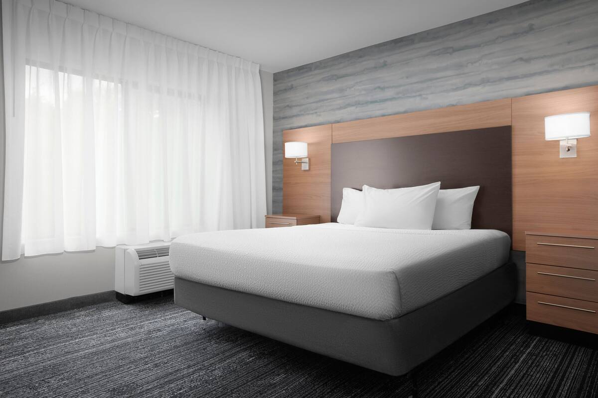 Une chambre d’hôtel moderne avec un grand lit, deux tables de chevet, une lampe fixée au mur et une fenêtre ornée de rideaux légers.