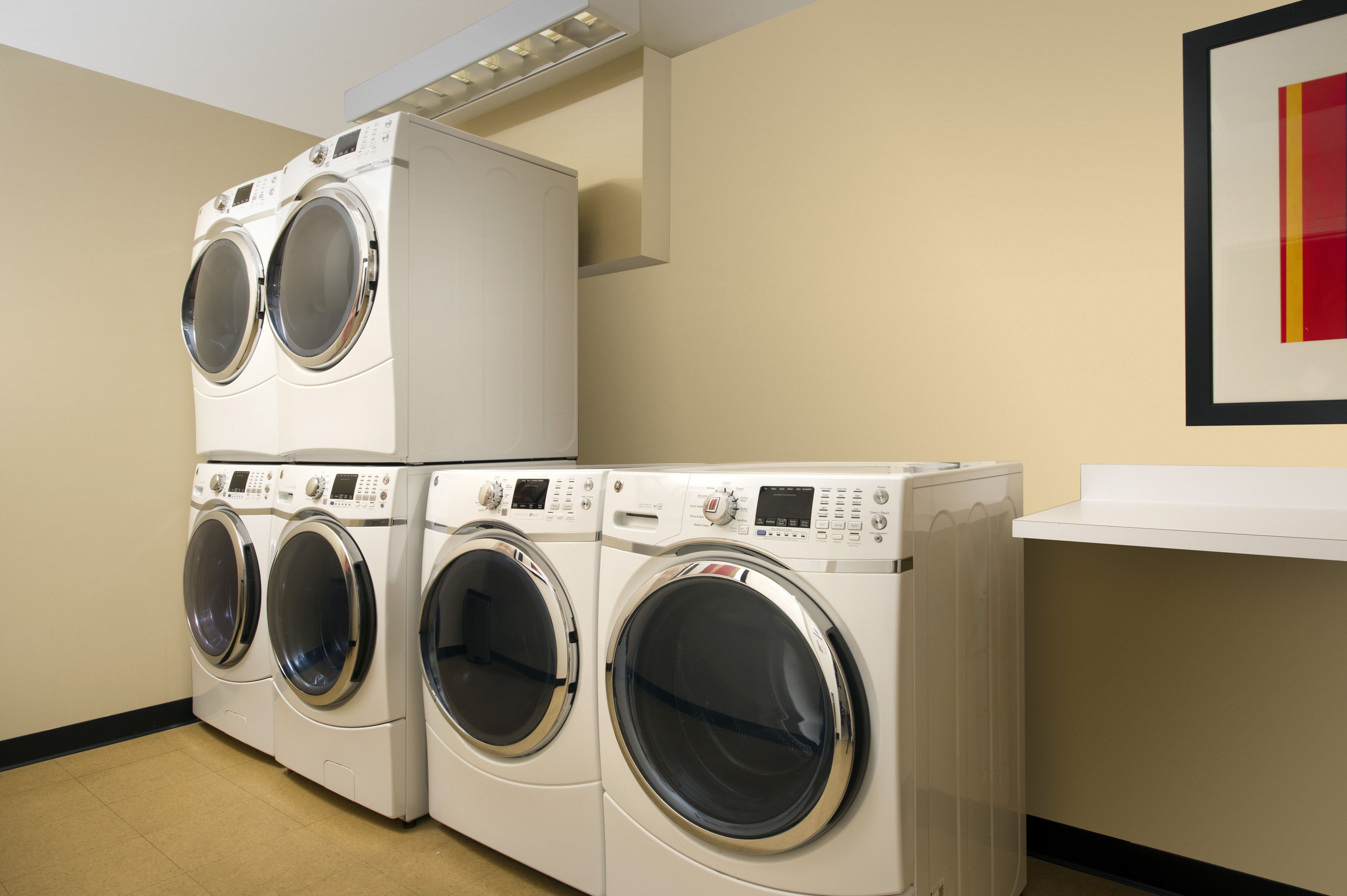 A laundry room with stacked front-loading washers and dryers, a white countertop, and a framed picture on the wall.