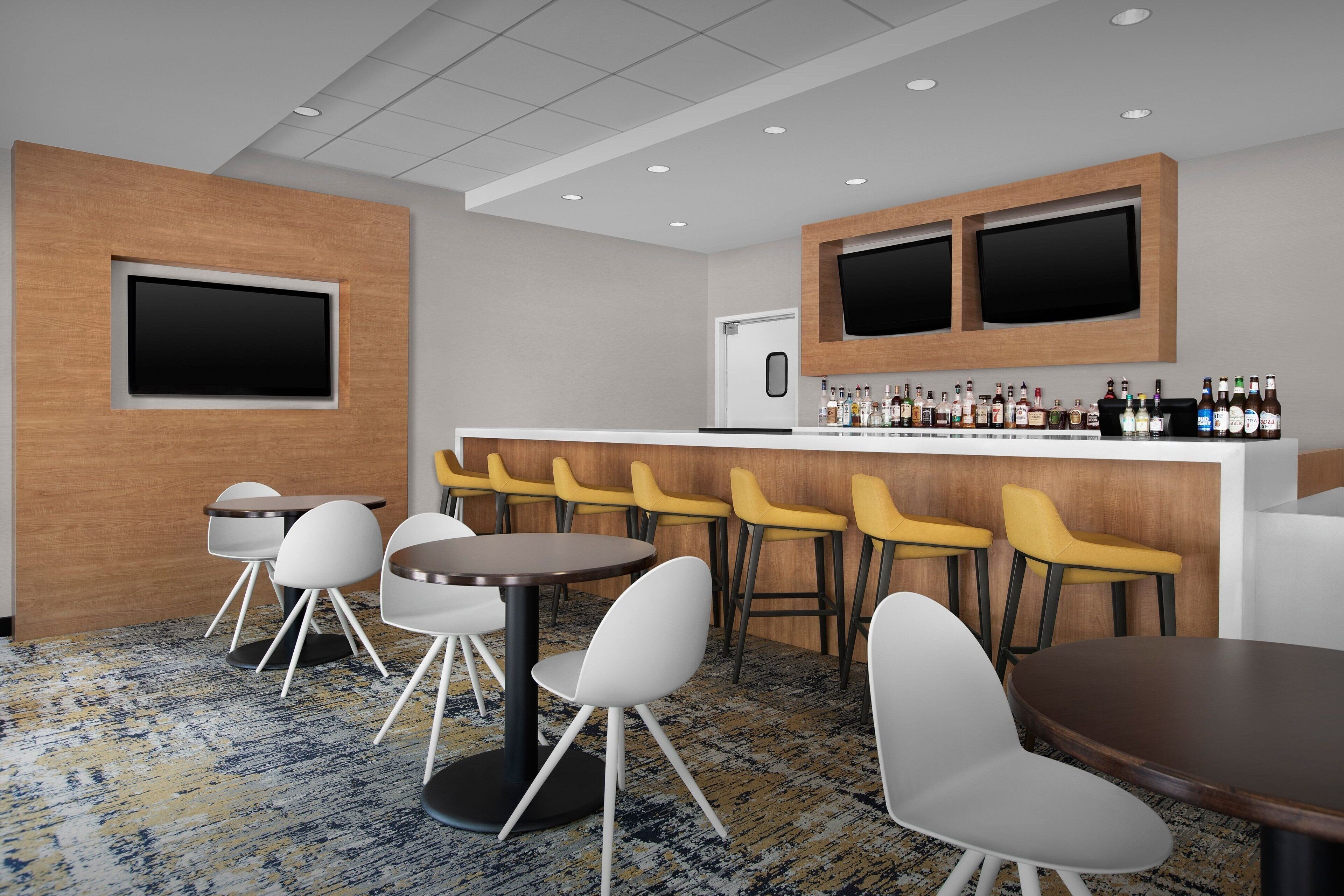 A modern bar area with multiple seating options, including round tables and bar stools, and a counter with a variety of beverages.