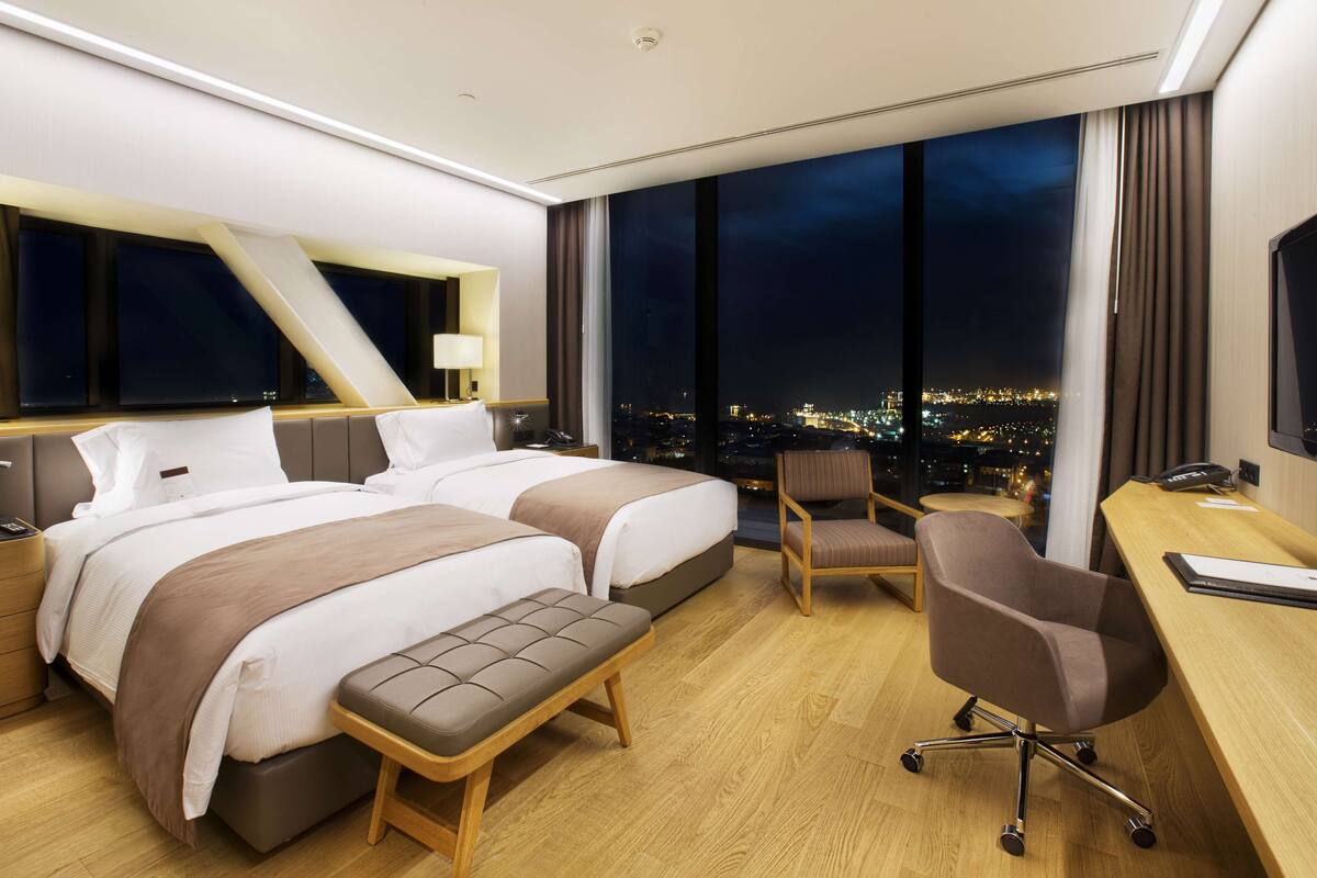 Una habitación de hotel moderna con una cama grande, un escritorio y una silla. Se ve la ciudad de noche a través de amplias ventanas.