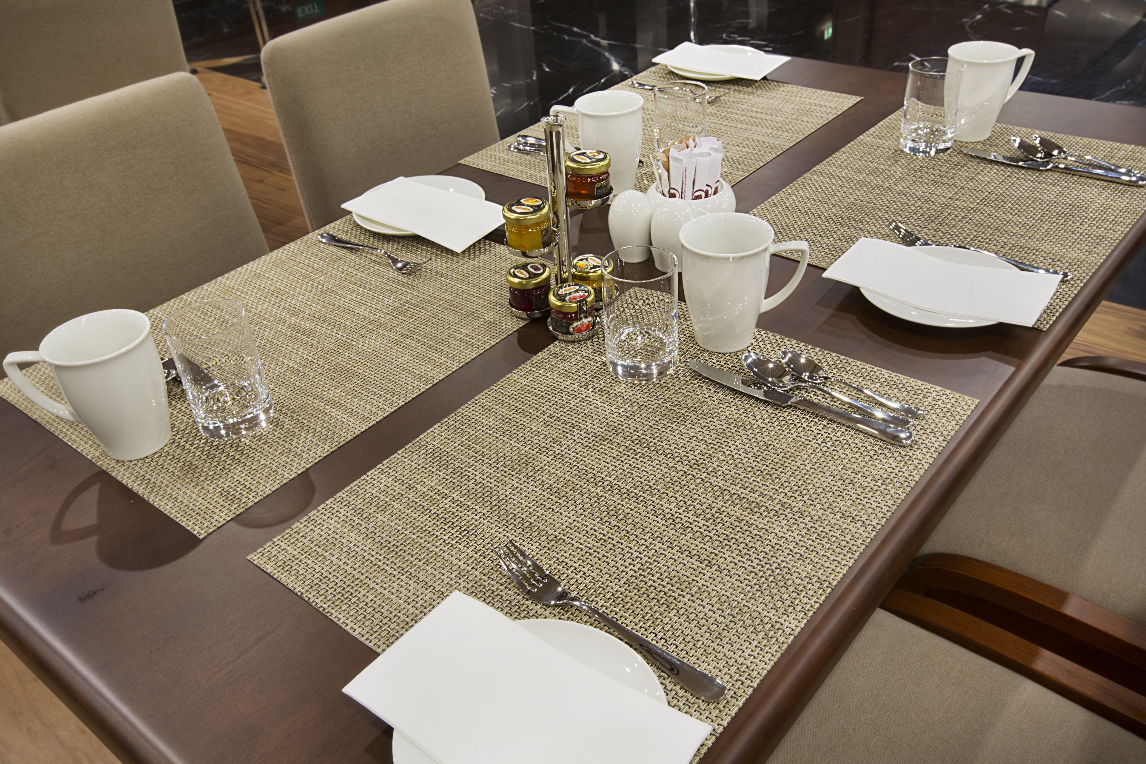 Mesa de comedor dispuesta con platos blancos, cubiertos y vasos transparentes, rodeada de sillas color beige.
