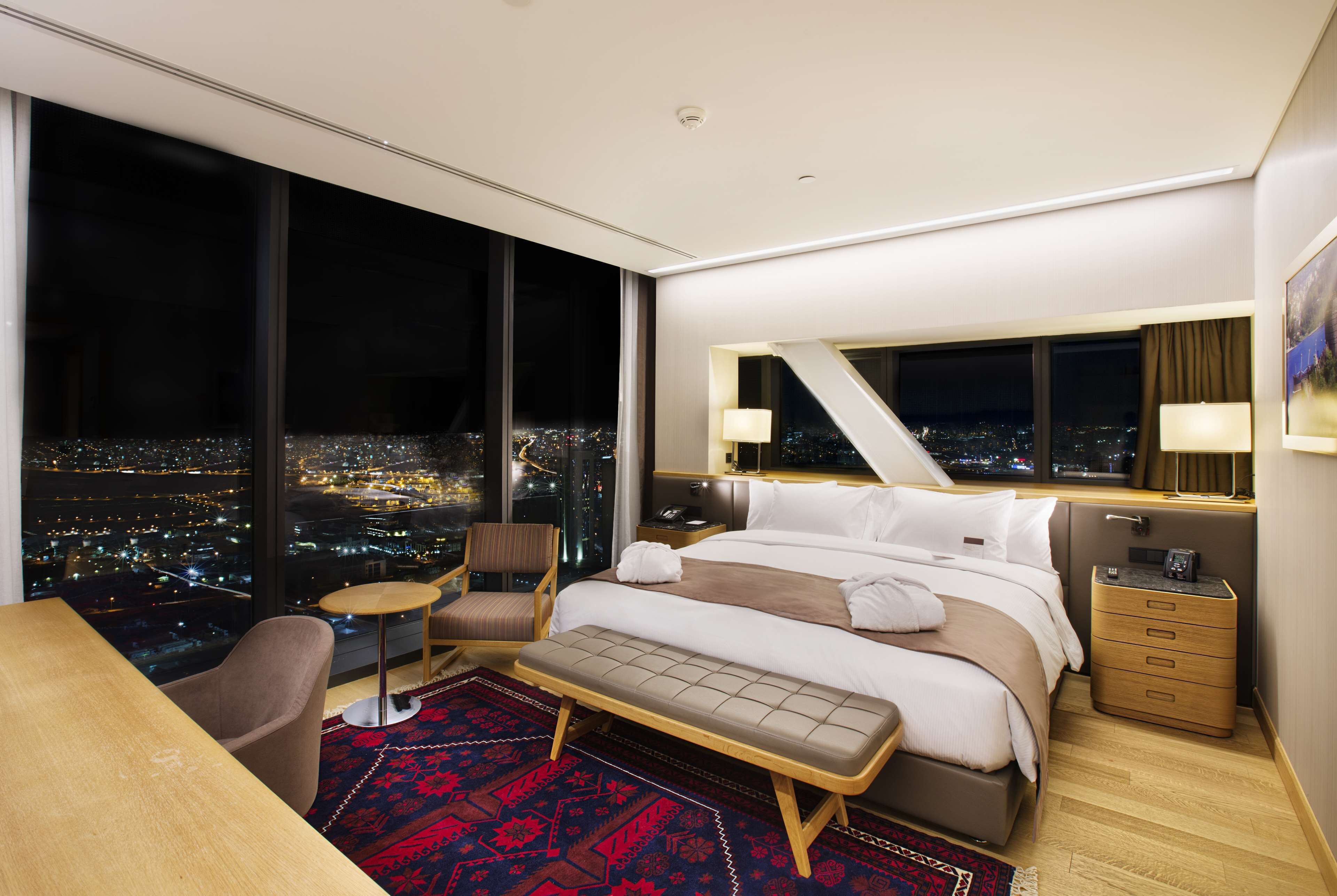 A modern hotel room with a large bed, a desk, a chair, and a view of a cityscape at night.