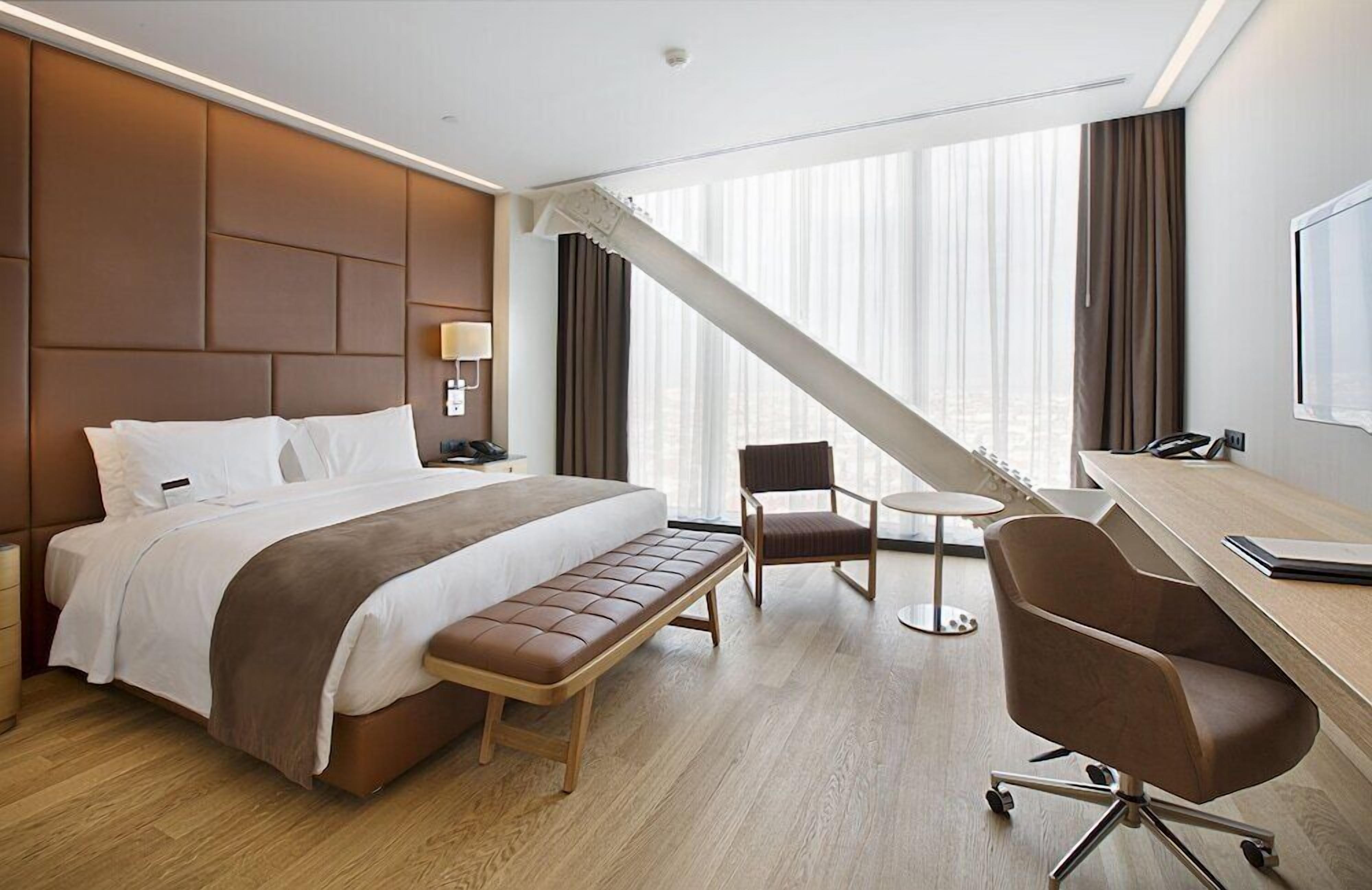 A hotel room with a bed, a desk, a chair, a bench, and a large window with curtains.