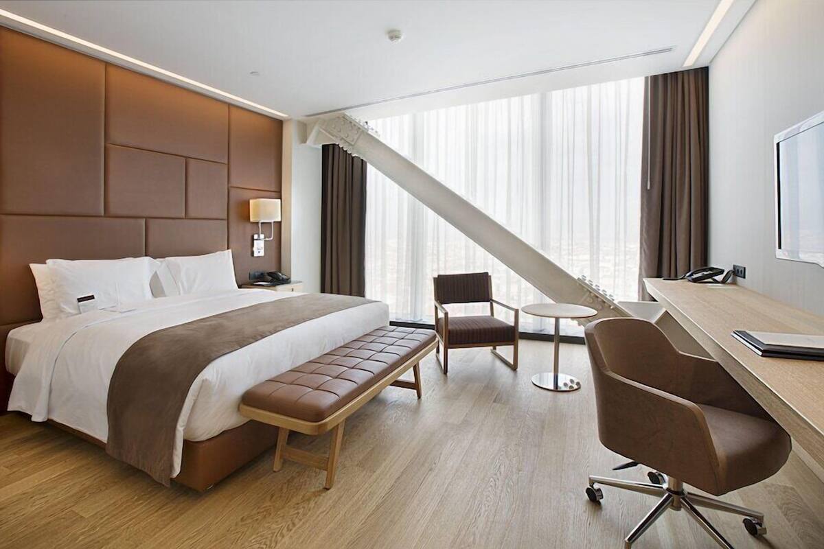 Habitación de hotel con cama, escritorio, silla, banco y un amplio ventanal con cortinas.