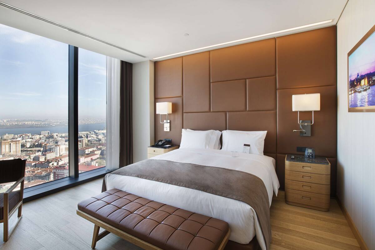 Una habitación de hotel moderna con una cama grande, un banco, una mesita de noche y vistas a la ciudad.