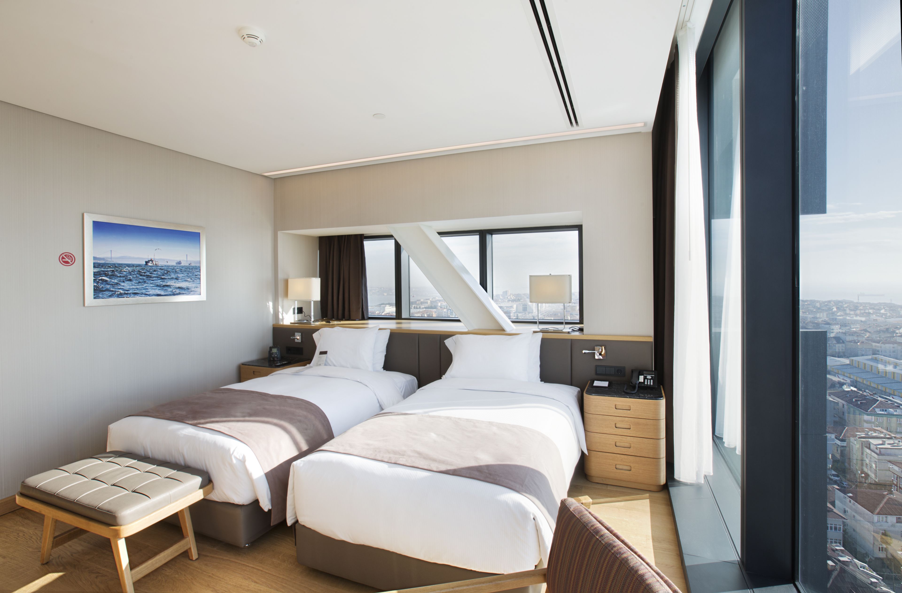 A modern hotel room with two beds, a small table, and a large window offering a city view.