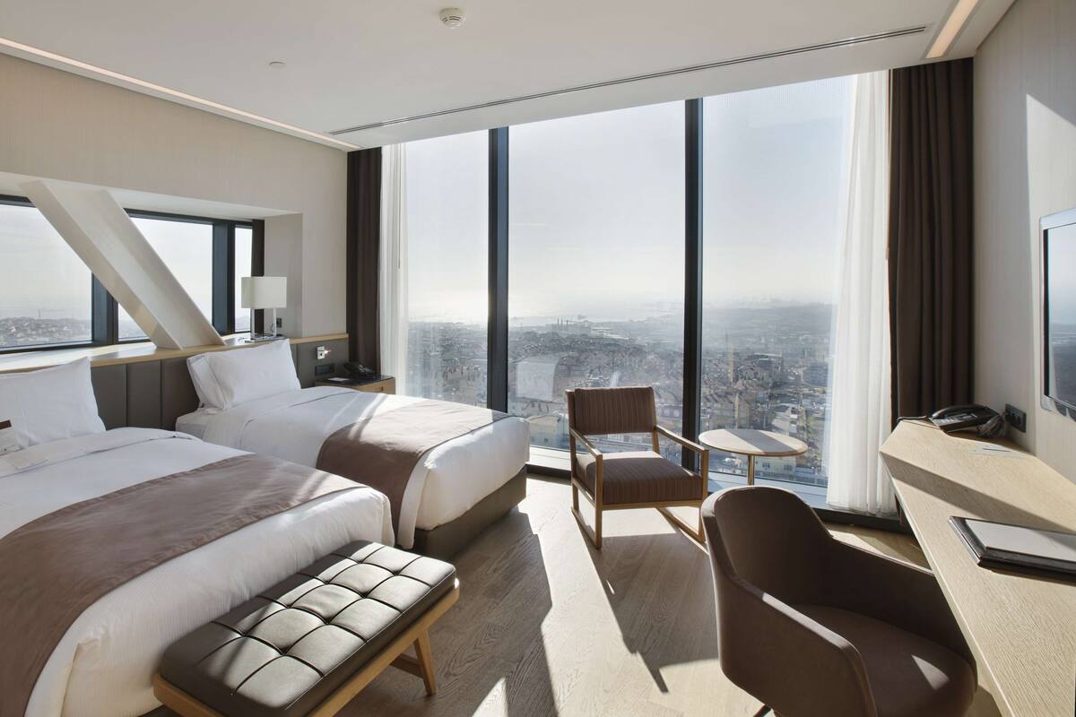 Una habitación de hotel moderna con dos camas, un rincón de descanso, un escritorio y grandes ventanas con vistas a la ciudad.
