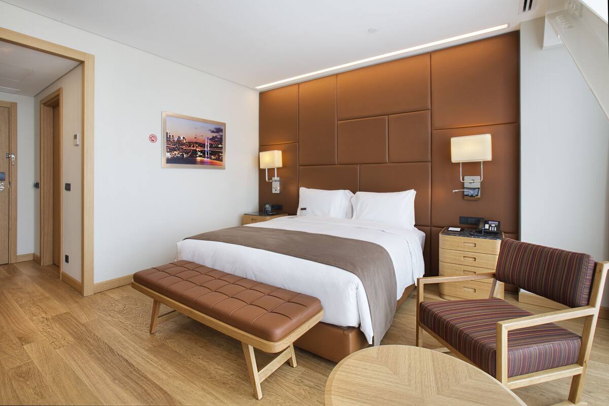 Habitación de hotel con una cama grande, un banco, una silla, una mesita de noche y una lámpara de pared.