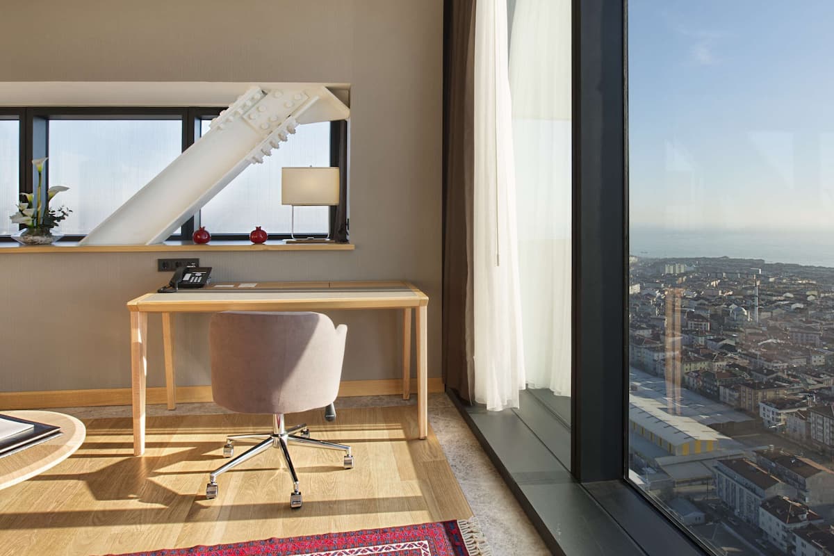 Una habitación de hotel moderna con un ventanal grande que ofrece vistas a la ciudad, un escritorio con una silla, una mesita con una granada y una planta decorativa.