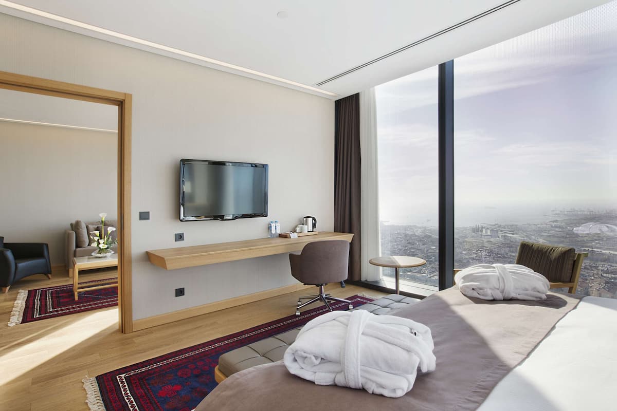 Una habitación de hotel moderna con una cama grande, un televisor montado en la pared, un escritorio pequeño y vistas de la ciudad a través de amplias ventanas.