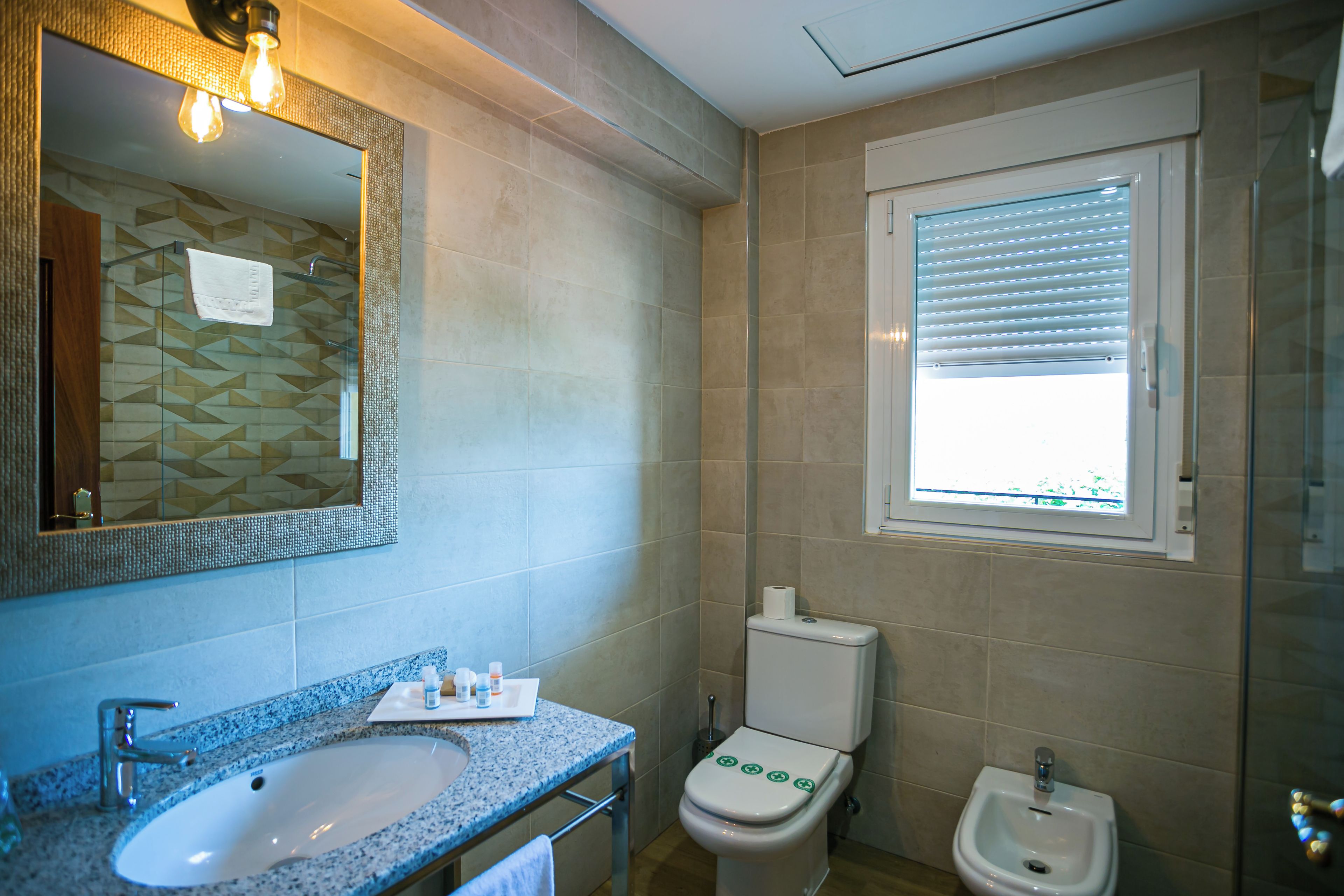 Une salle de bain équipée d’un lavabo, d’un miroir, de toilettes, d’un bidet et d’une fenêtre avec des stores.