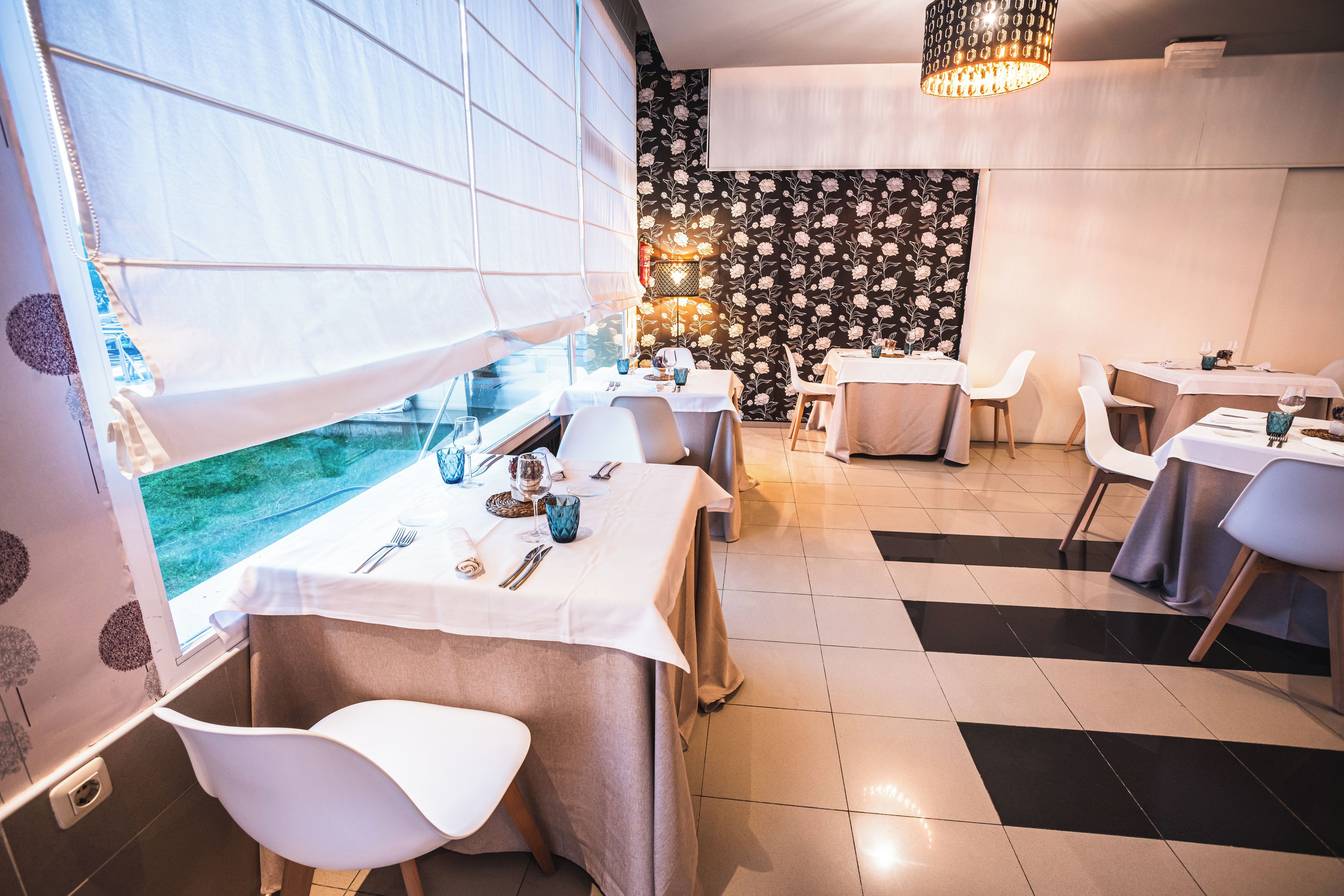 A restaurant with tables set for dining, a patterned wall, and a window with blinds.
